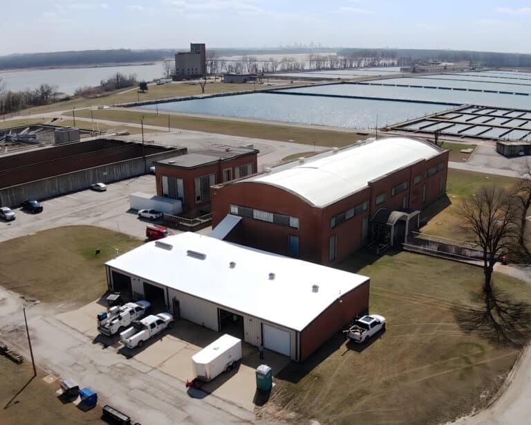 Aerial view of a water treatment plant with a curved white roof, a flat-roofed garage building, and settling basins by a river.