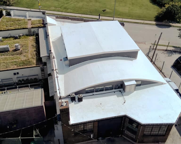 Aerial view of a brick building with a white flat roof, curved raised section, and a neighboring green roof.